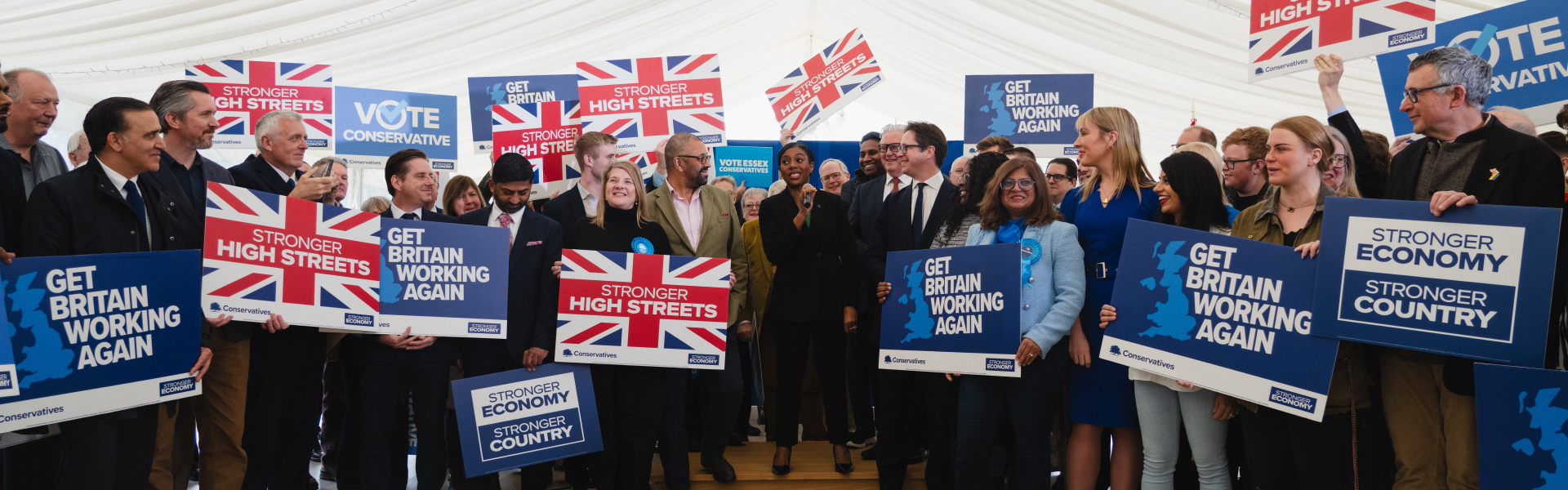 A Conservative Party campaign rally in Essex with Leader of the Conservative Party Kemi Badenoch MP and Shadow Secretary of State for Housing, Communities and Local Government Sir James Cleverly MP, surrounded by activists holding signs reading “Get Britain Working Again”, “Stronger High Streets”, “Vote Conservative”, and “Stronger Economy, Stronger Country”.